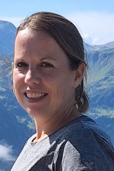 Julia Lock Julia Lock - Porträt. Man sieht nur ihr Gesicht, ihre etwa schulterlangen Haare sind zu einem Zopf gebunden. Im Hintergrund sieht man Berge.