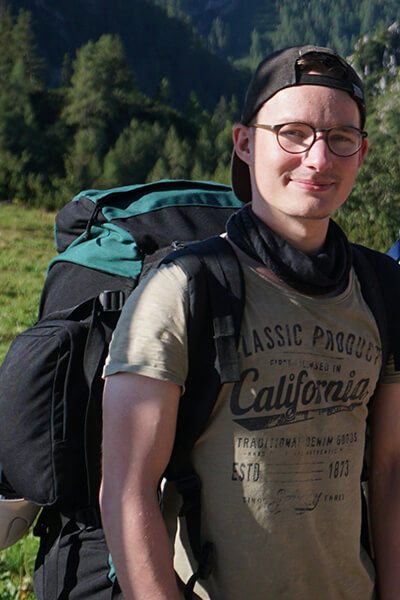 Julius Krastel Junger Mann mit Wanderrucksack, Brille und Cappi. Er steht in der Natur.