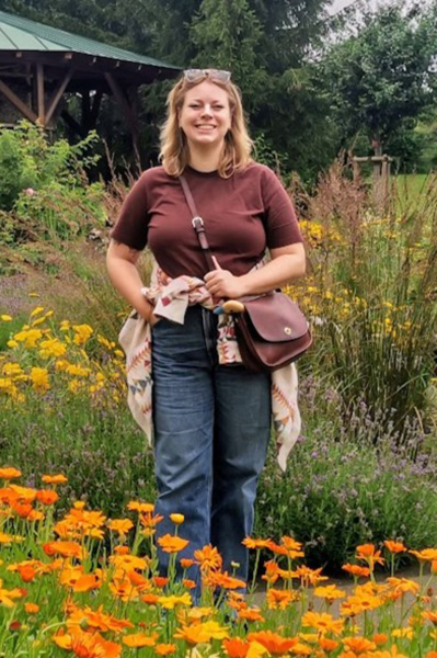 Zoë Richardson Porträt einer Frau, die in einem Park steht, man sieht viele bunte Blumen, sie trägt eine lange Hose und ein T-Shirt.