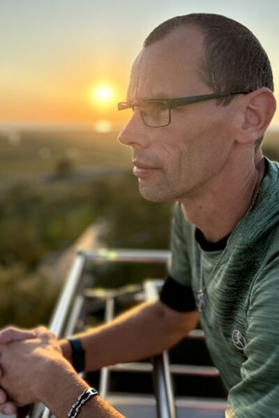 Mann - von der Seite fotografiert - lehnt auf einem Balkongeländer, im Hintergrund sieht man die untergehende Sonne. 