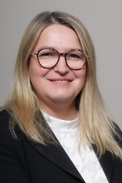Porträt einer Frau mit blonden langen Haaren, schwarzem Sakko und Brille