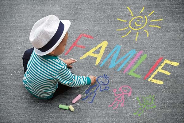 Kreidezeichnung "Familie" Kleinkind sitzt mit Kreide auf der Straße. Man sieht Strichmännchen und die Schrift "Familie". Quelle: Adobe Stock