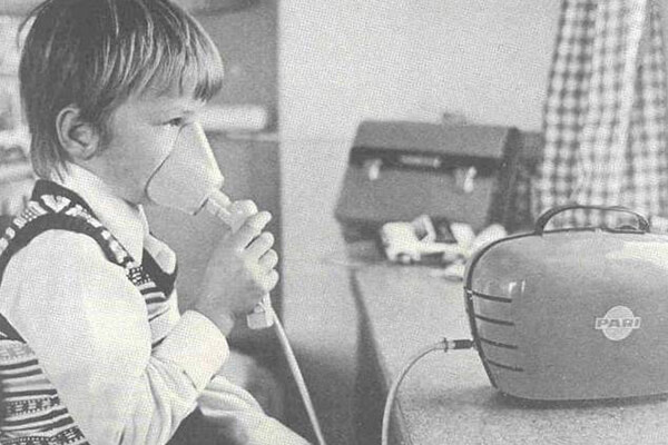 Inhalation in den 1970er Jahren Schwarzweiß Foto von einem Jungen, der mit einem größeren Inhalationsgerät inhaliert. Man erkennt, dass das Bild schon um die 50 Jahre alt sein muss, da die Kleidung und die Möbel darauf hinweisen.