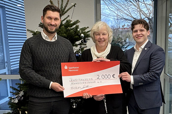 Zwei Männer (links und rechts außen) und eine Frau (Mitte) stehen vor einem Tannenbaum. Sie alle halten einen symbolischen Spendenscheck in der Hand, auf dem steht: 2.000 Euro.
