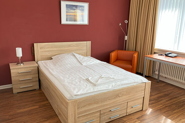 Ein Zimmer im Haus Schutzengel mit Bett, roter Wand, orangenem Sessel, Leselampe und Fensterfront. 