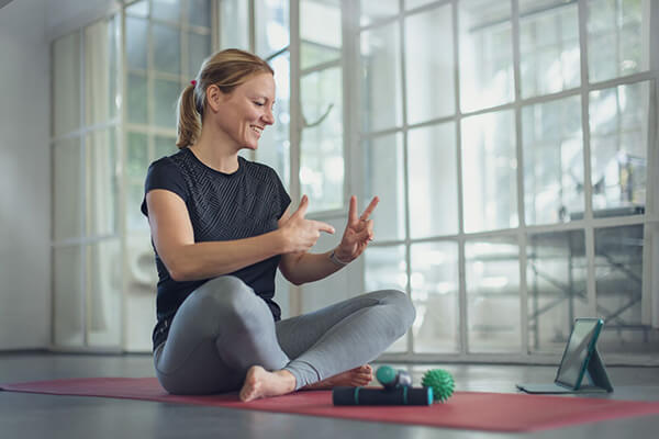 Sportübung beim Online-Sport Frau in Sportkleidung sitzt auf einer Yoga-Matte und macht eine Übung vor einem Tablet vor.